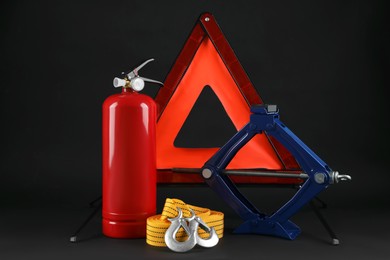 Photo of Emergency warning triangle, red fire extinguisher, towing strap and scissor jack on black background. Car safety