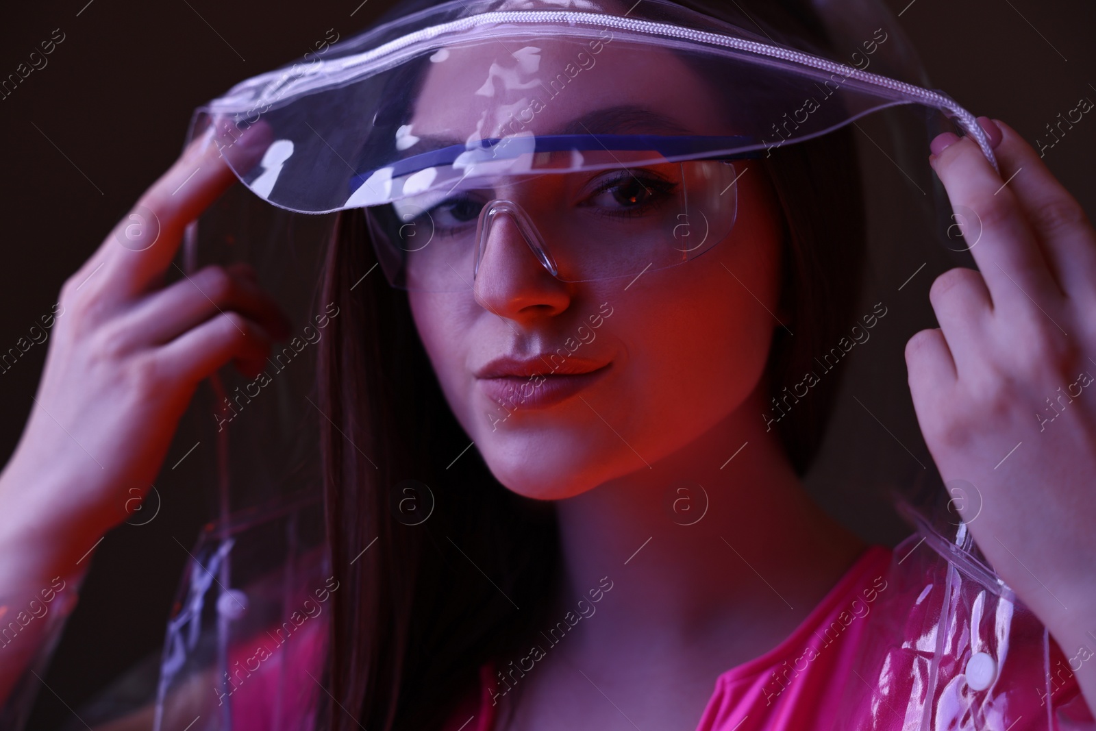 Photo of Fashionable portrait of beautiful woman wearing transparent coat and glasses on dark background in neon lights, closeup