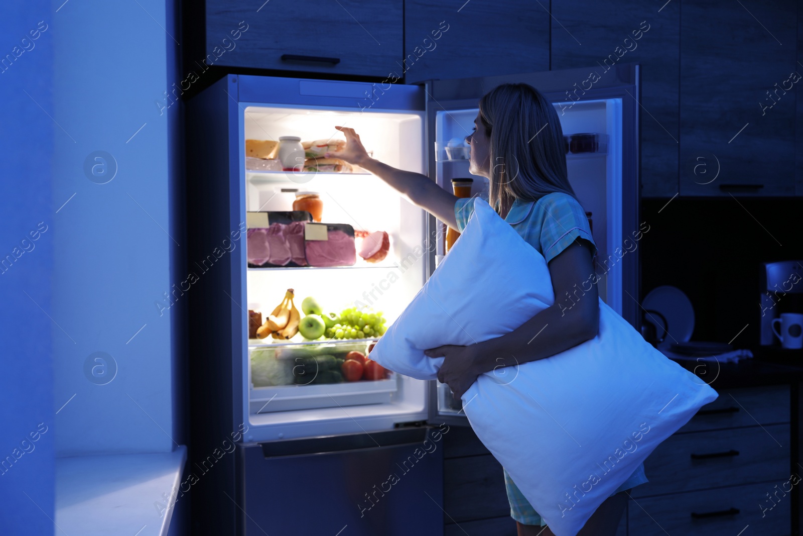 Photo of Woman with pillow looking into refrigerator in kitchen at night