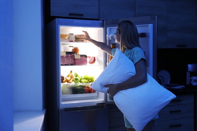 Woman with pillow looking into refrigerator in kitchen at night