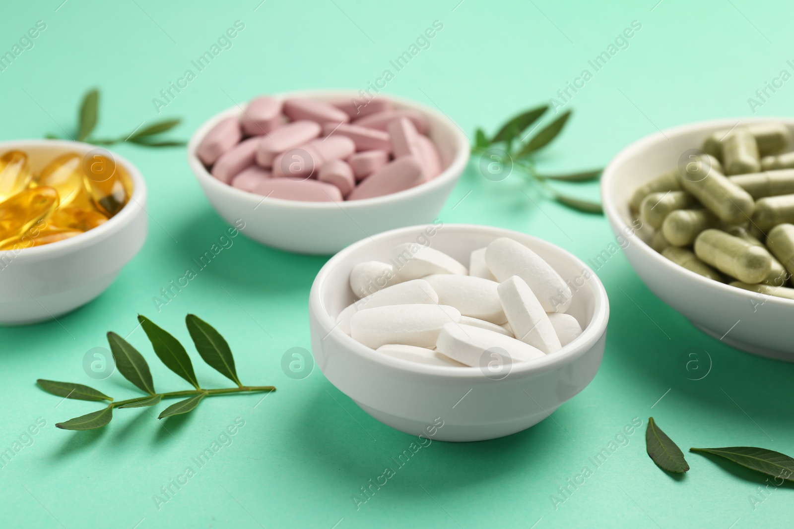 Photo of Different vitamin capsules in bowls and leaves on turquoise background, closeup