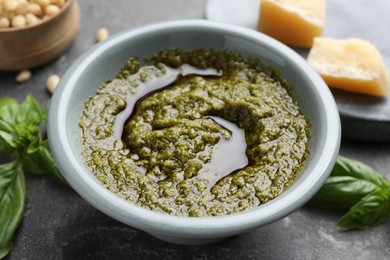 Photo of Tasty pesto sauce in bowl, basil, cheese and pine nuts on grey table, closeup