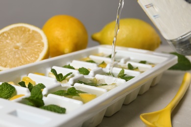 Pouring water into ice cube tray with mint and lemon on table, closeup