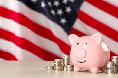 Photo of Piggy bank and coins on white wooden table against American flag, space for text