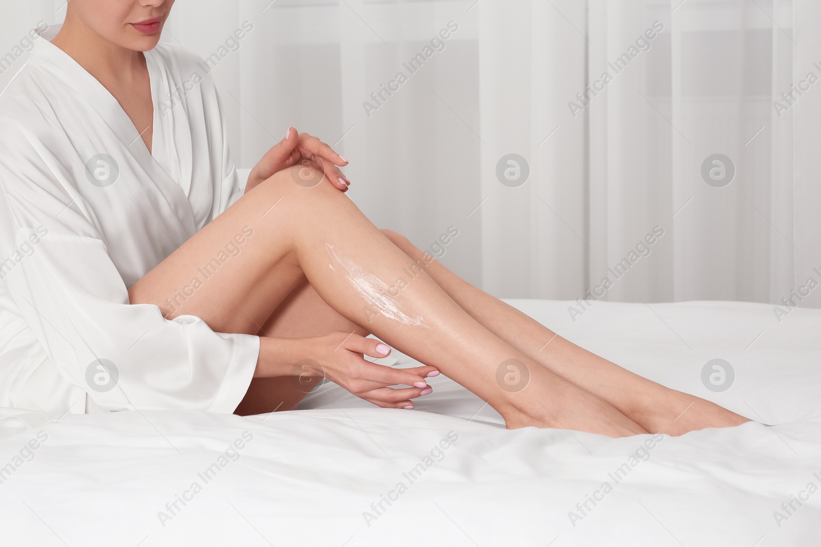 Photo of Woman applying body cream onto her smooth legs on bed indoors, closeup