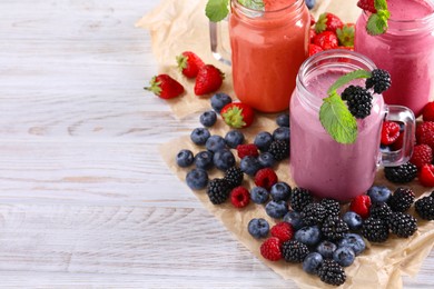 Photo of Mason jars of different berry smoothies and fresh ingredients on white wooden table. Space for text