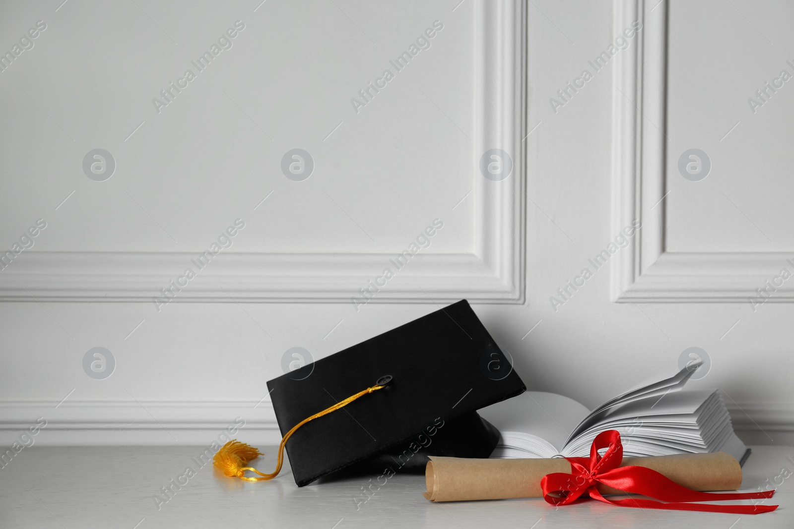 Photo of Graduation hat, book and diploma on floor near white wall, space for text