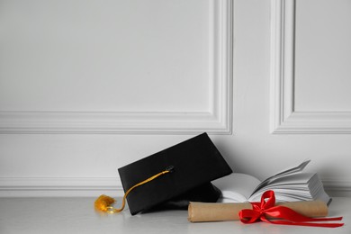 Graduation hat, book and diploma on floor near white wall, space for text