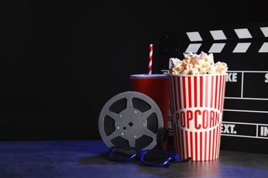 Clapperboard, reel and popcorn on table against dark background, space for text. Watching cinema