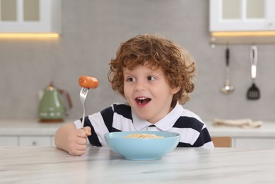Photo of Cute little boy holding fork with sausage and bowl of pasta at table in kitchen