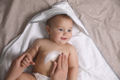 Photo of Mother applying moisturizing cream onto her little baby's skin on bed, closeup