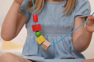 Photo of Little girl playing with wooden pieces and string for threading activity on light background, closeup. Child's toy