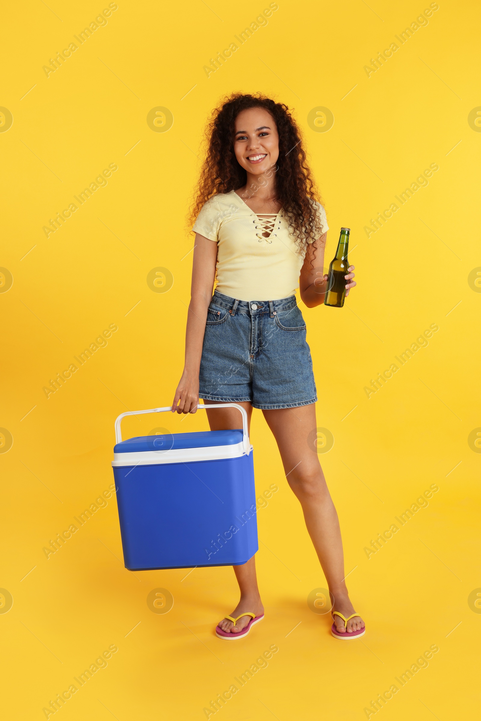 Photo of Happy young African American woman with cool box and bottle of beer on yellow background