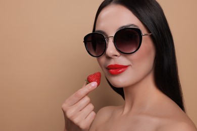 Photo of Attractive woman in fashionable sunglasses holding strawberry against beige background. Space for text