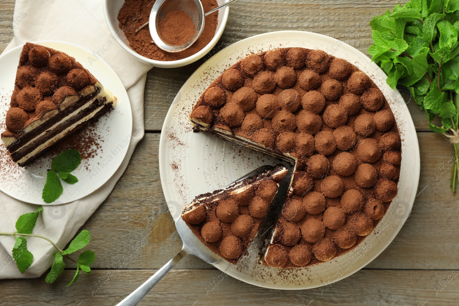 Photo of Delicious tiramisu cake, cocoa powder, server and mint leaves on wooden table, flat lay
