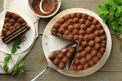 Photo of Delicious tiramisu cake, cocoa powder, server and mint leaves on wooden table, flat lay
