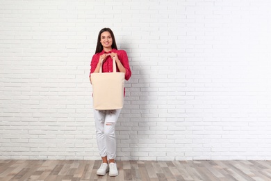 Beautiful young woman with stylish blank eco bag near brick wall, space for text