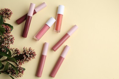 Photo of Different lip glosses and flowers on pale yellow background, flat lay. Space for text