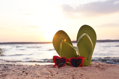 Stylish flip flops with sunglasses on sand near sea, space for text. Beach accessories