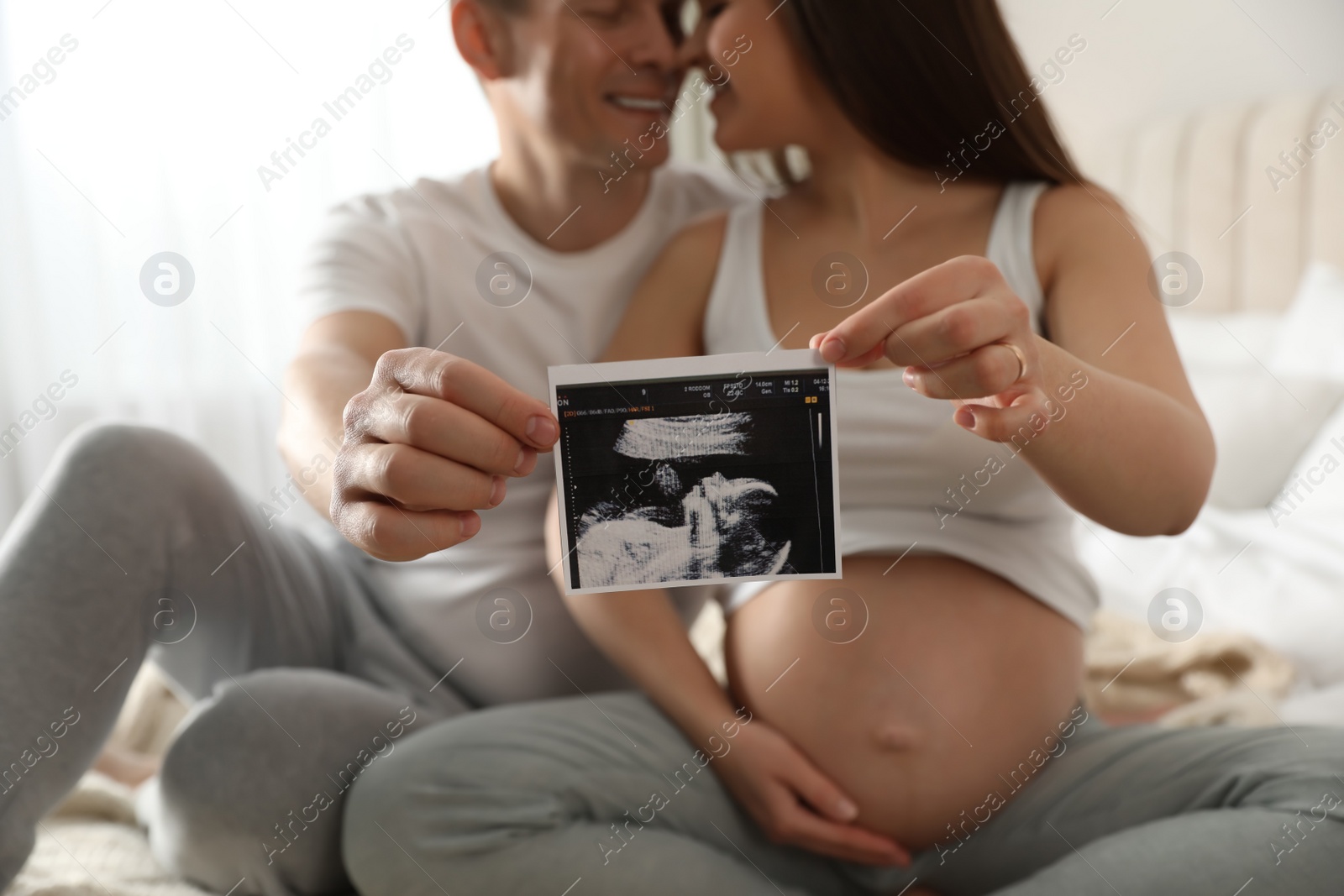 Photo of Young pregnant woman with her husband in bedroom, focus on ultrasound picture of baby