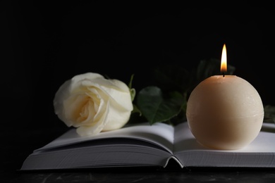 Photo of Burning candle, white rose and book on table in darkness, closeup. Funeral symbol