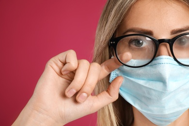 Photo of Woman wiping foggy glasses caused by wearing disposable mask on pink background, closeup. Protective measure during coronavirus pandemic