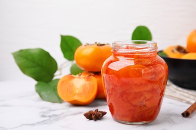 Jar of tasty persimmon jam and ingredients on white marble table. Space for text