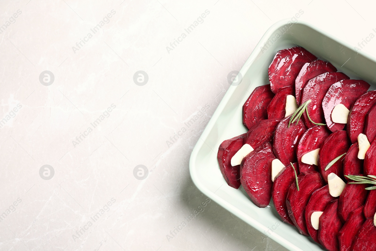 Photo of Baking dish with raw beetroot slices, garlic and rosemary on light marble table, top view. Space for text