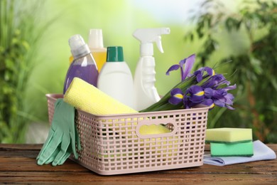 Photo of Spring cleaning. Plastic basket with detergents, supplies and beautiful flowers on wooden table outdoors