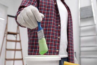 Photo of Man dipping brush into bucket of green paint indoors, closeup