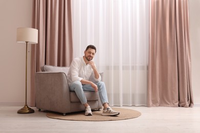Happy man resting on armchair near window with beautiful curtains at home. Space for text
