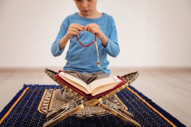 Little Muslim boy with misbaha and Koran praying on rug indoors, closeup