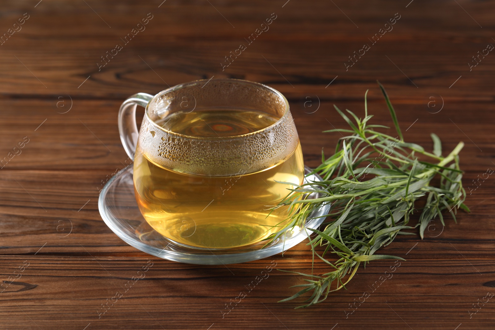 Photo of Cup of homemade herbal tea and fresh tarragon leaves on wooden table