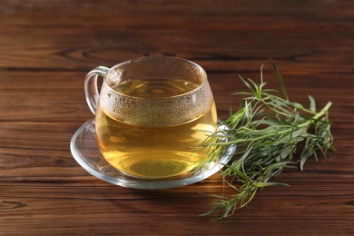 Photo of Cup of homemade herbal tea and fresh tarragon leaves on wooden table