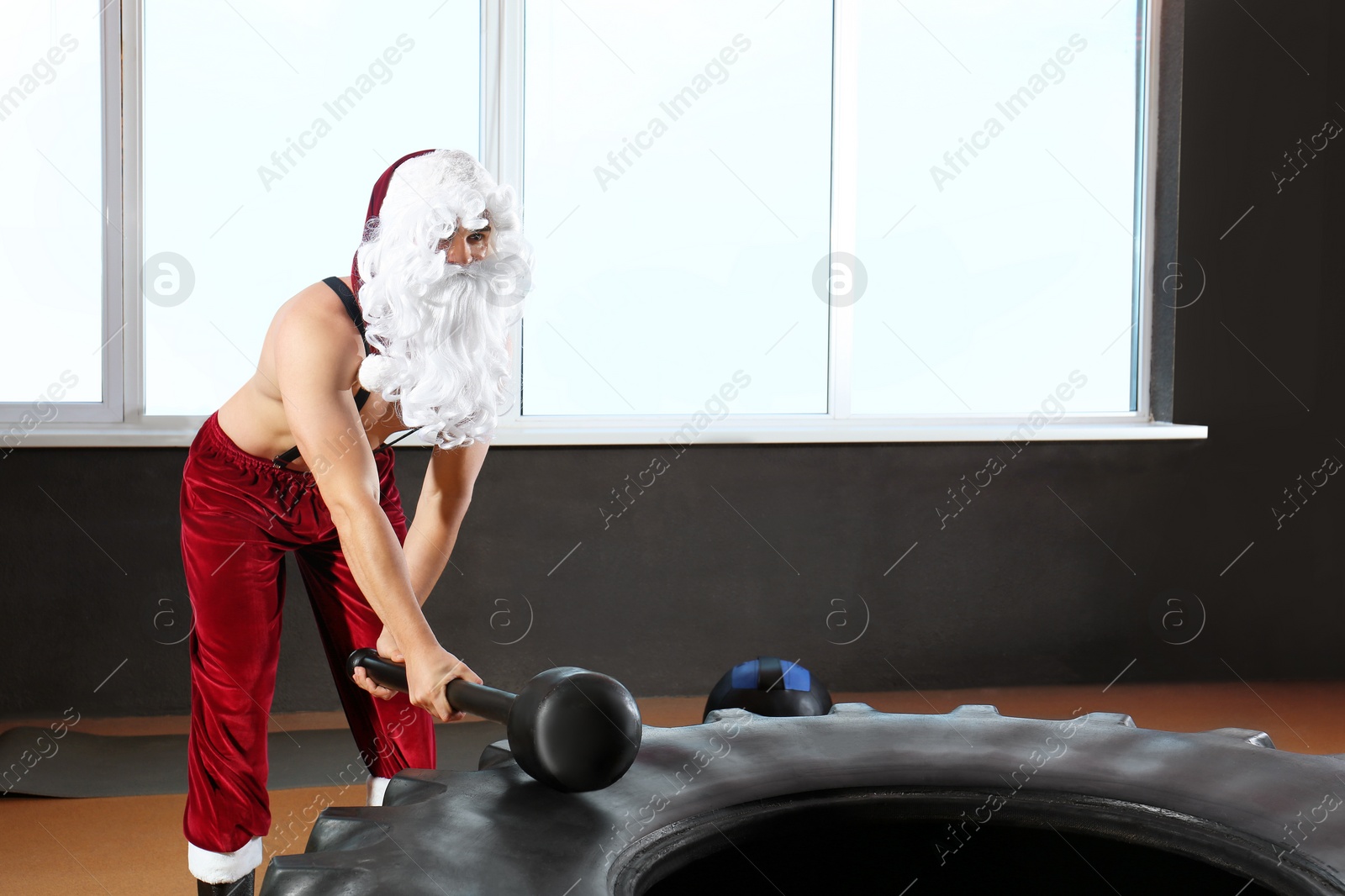 Photo of Young Santa Claus hitting heavy tire with hammer in modern gym