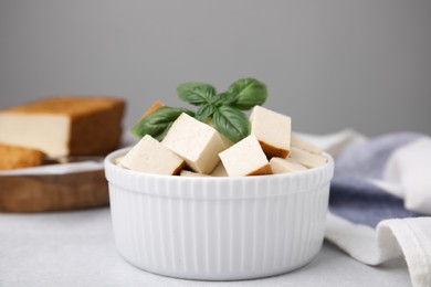 Photo of Bowl of smoked tofu cubes with basil on white table, closeup