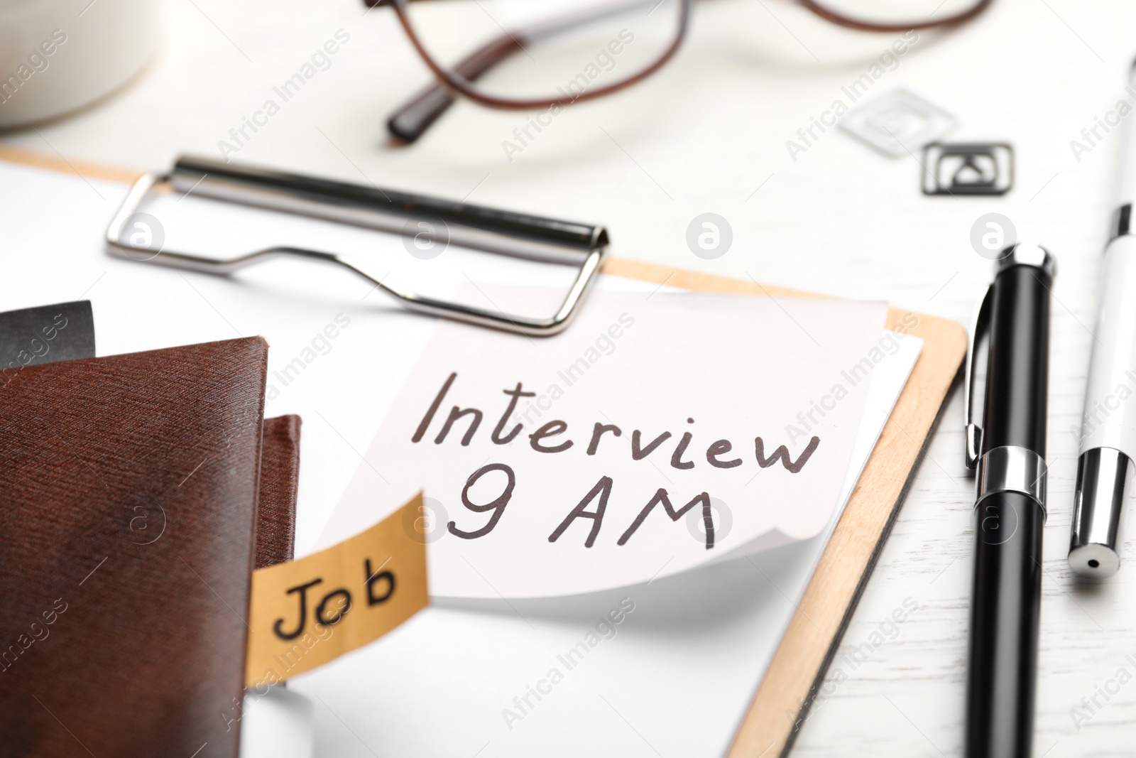 Photo of Clipboard with reminder note about job interview and stationery on table, closeup