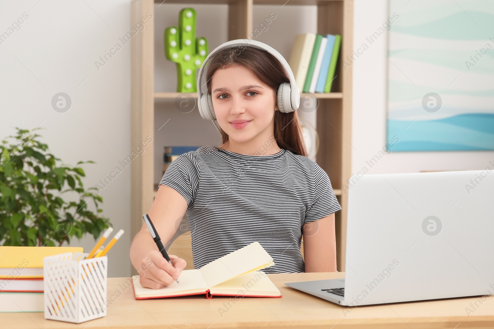 Photo of Cute girl using laptop and headphones at desk in room. Home workplace