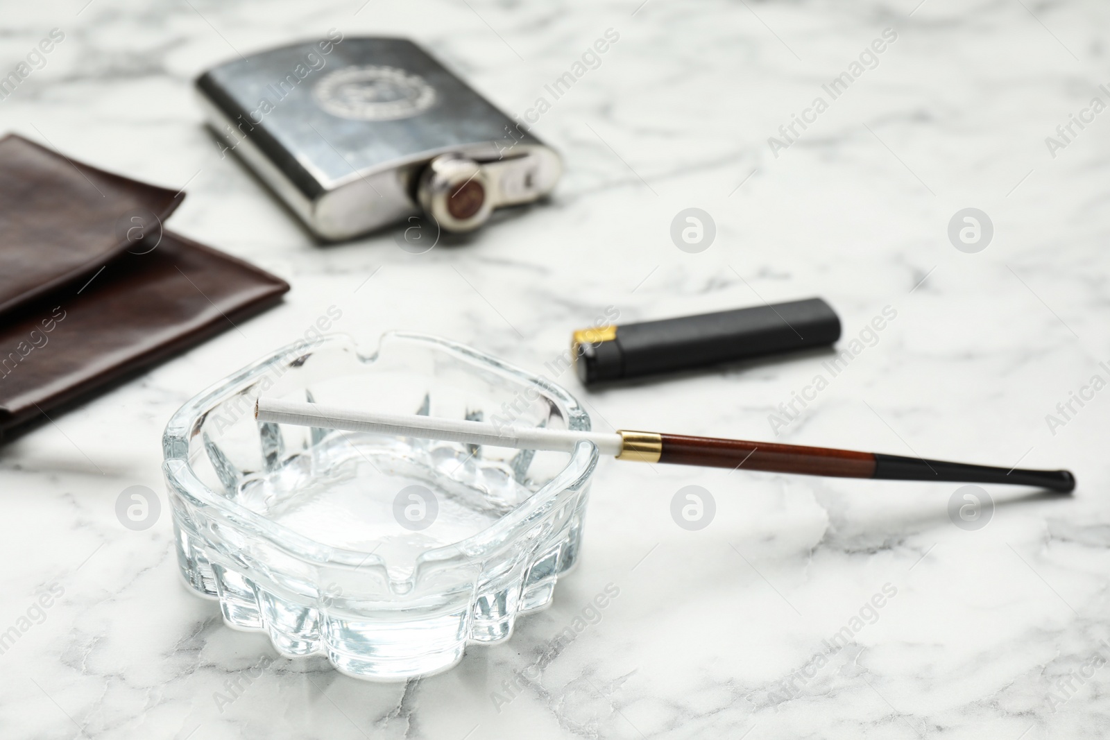 Photo of Glass ashtray with long cigarettes holder, clutch, pocket flask and lighter on white marble table