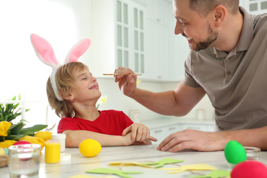 Photo of Happy son wearing bunny ears headband and his father having fun while at table in kitchen with Easter eggs