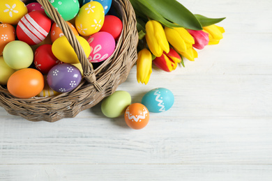 Photo of Colorful Easter eggs in basket and tulips on white wooden table, flat lay. Space for text