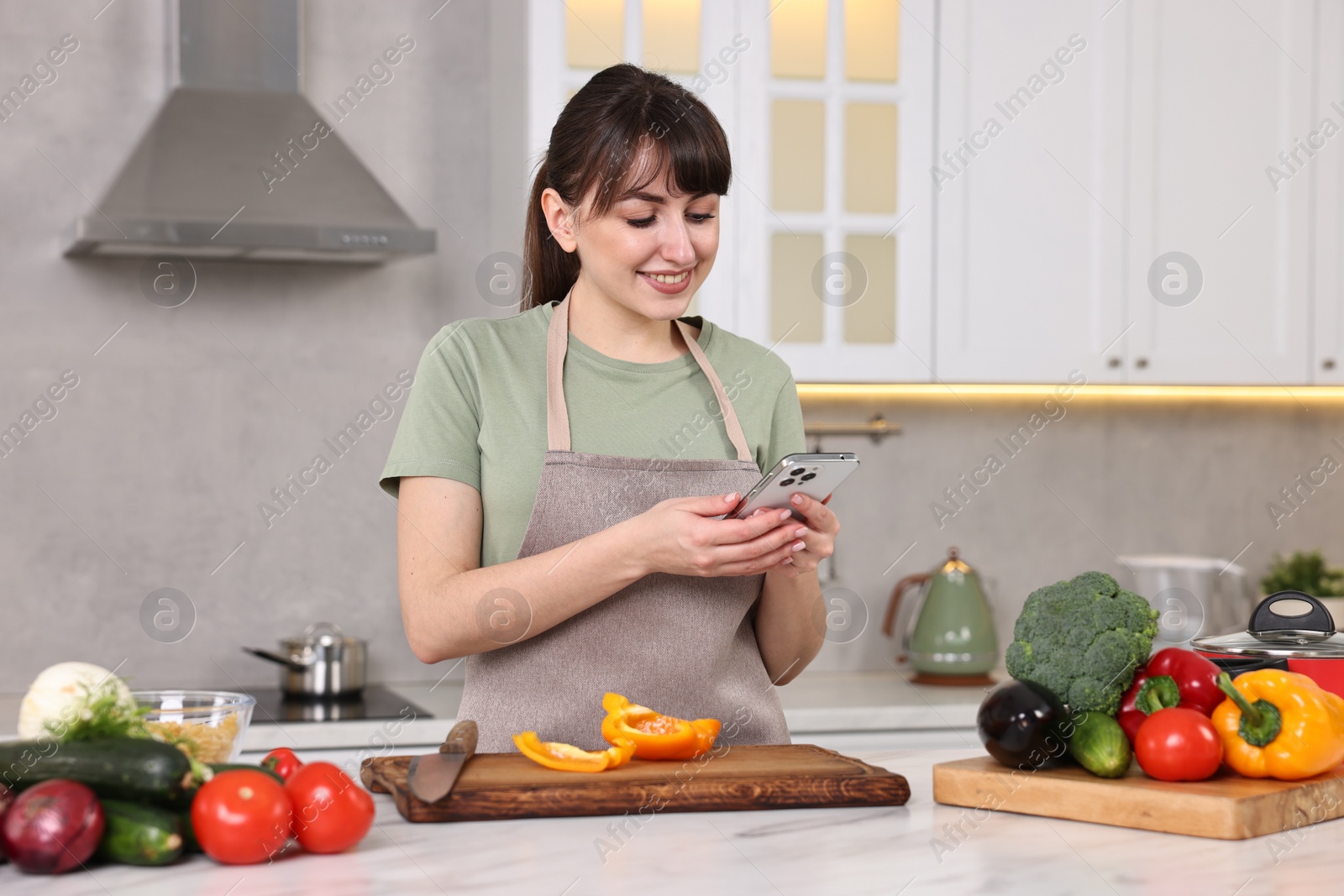 Photo of Happy young housewife using smartphone while cooking at white marble table in kitchen