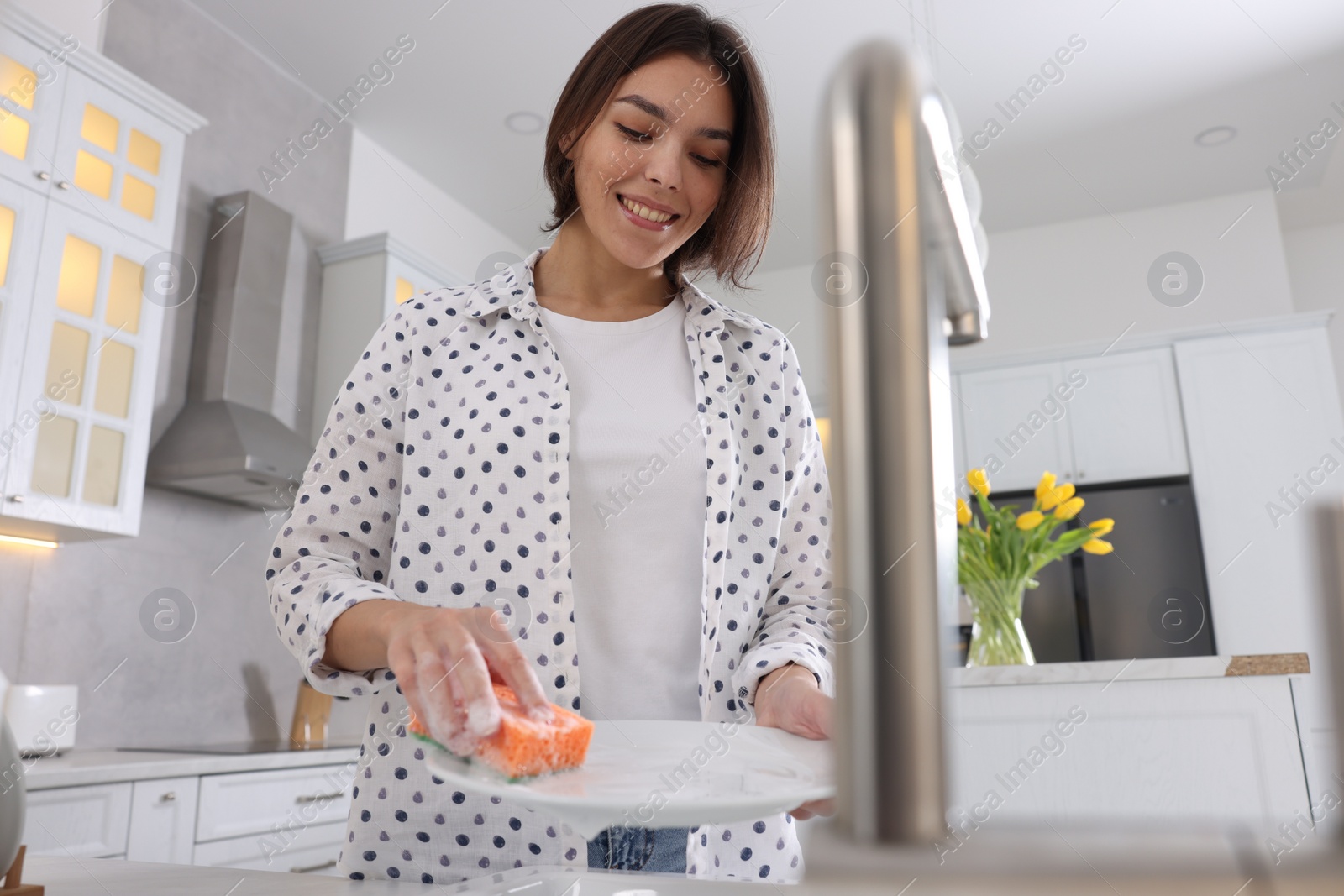 Photo of Happy young woman washing plate above sink in modern kitchen