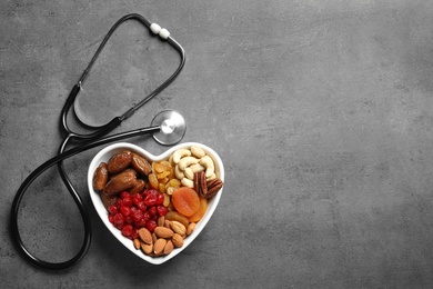 Photo of Heart shaped bowl with nuts and dried fruits near stethoscope on grey background, top view. Space for text