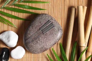 Stones with acupuncture needles and palm leaves on bamboo mat, flat lay