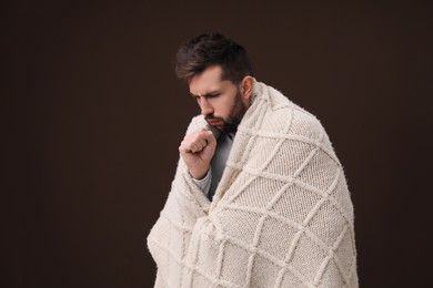Photo of Man wrapped in blanket coughing on brown background. Cold symptoms