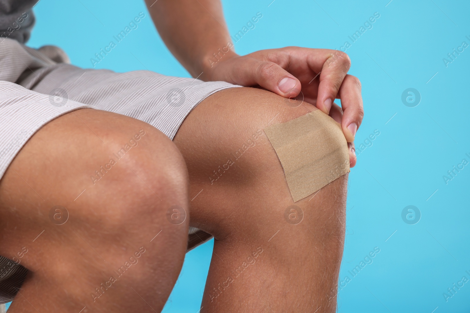 Photo of Man putting sticking plaster onto knee on light blue background, closeup