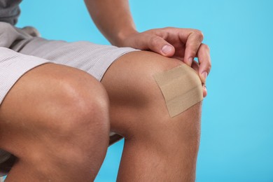 Photo of Man putting sticking plaster onto knee on light blue background, closeup