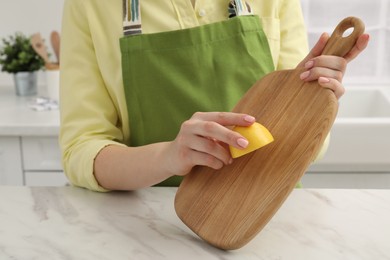 Photo of Woman rubbing wooden cutting board with lemon at white table in kitchen, closeup