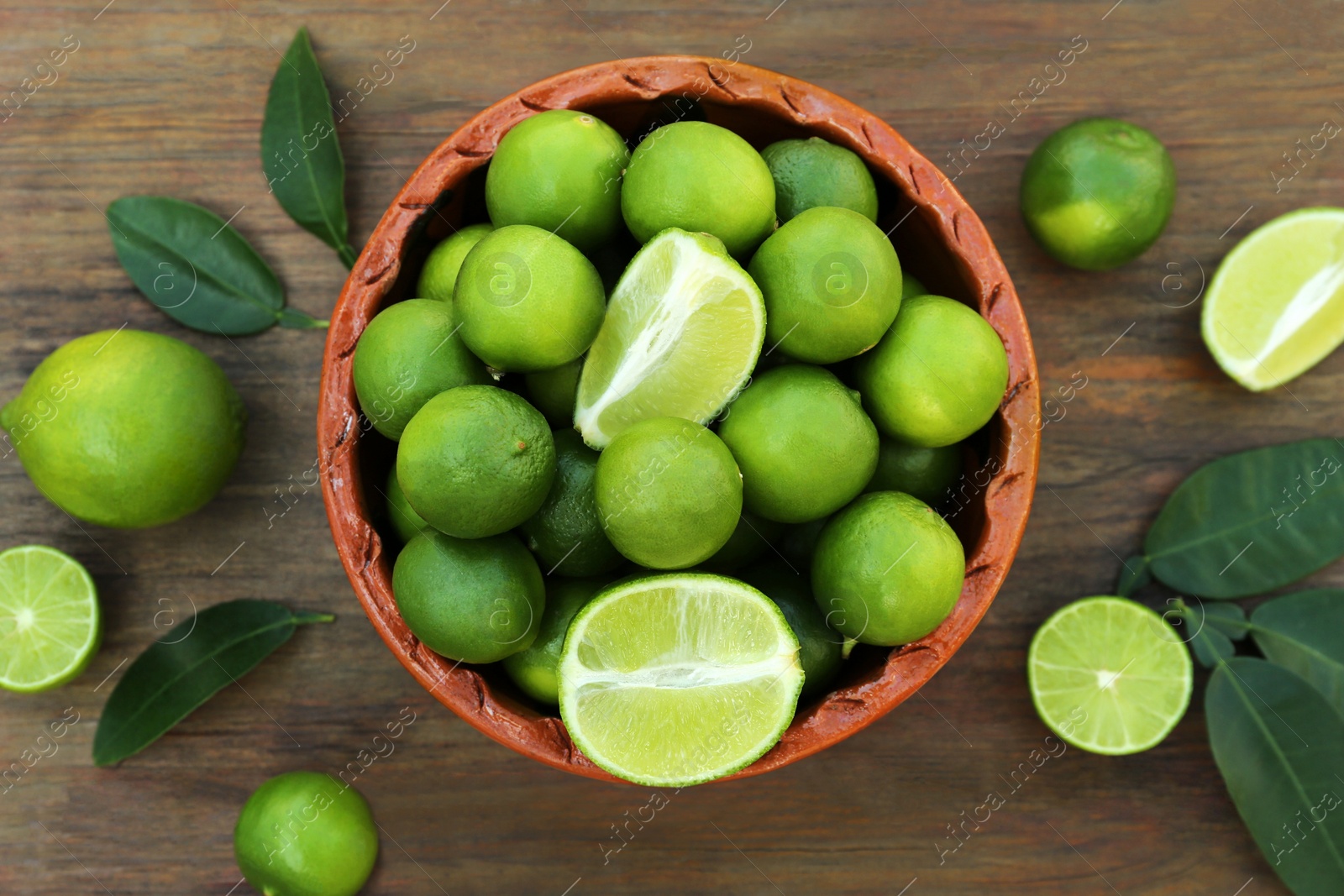 Photo of Whole and cut fresh ripe limes in bowl on wooden table, flat lay